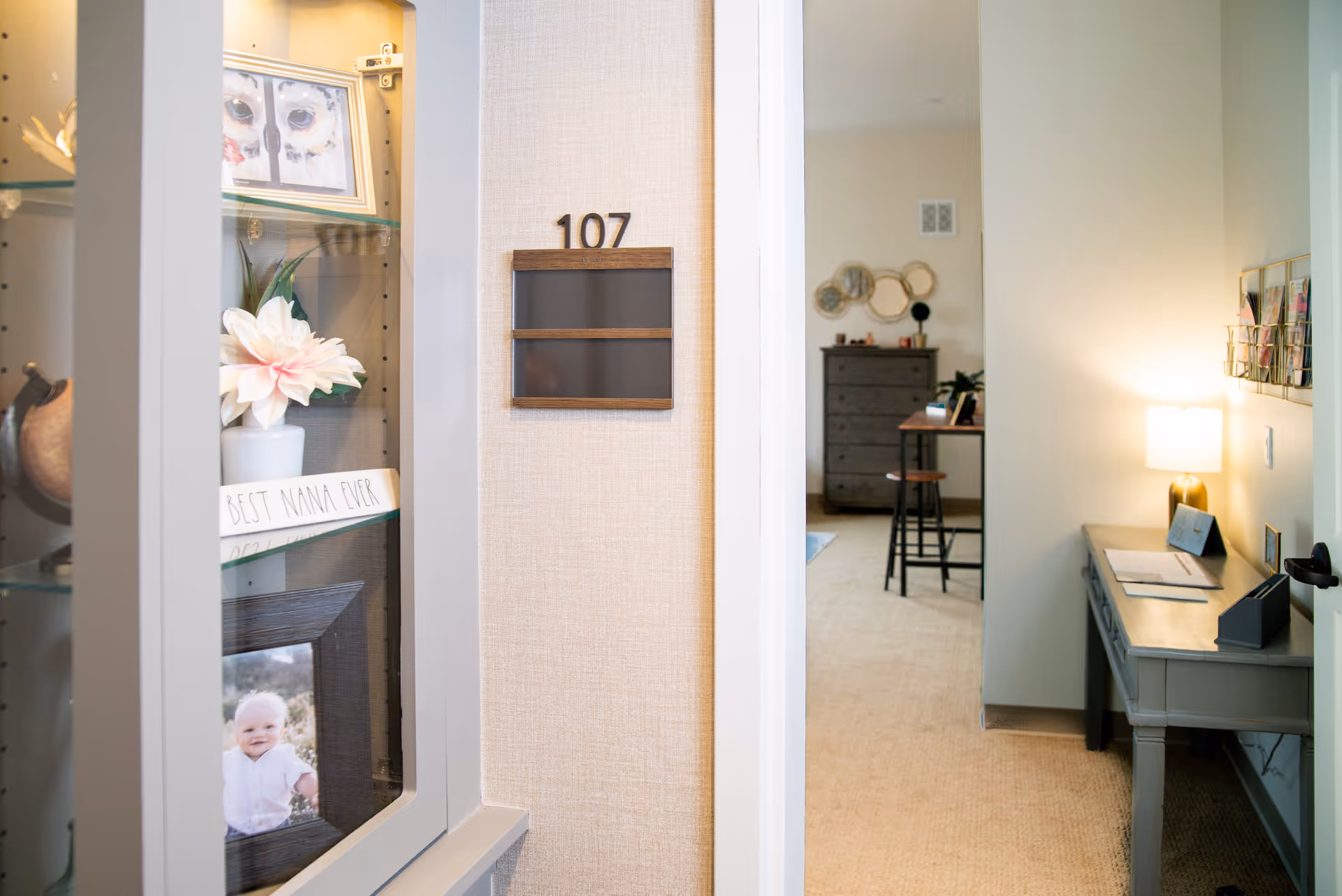 View into a senior living facility room numbered 107, showing a hallway with a small desk and lamp on the right, and a display cabinet on the left containing a flower, framed photos, and a sign that reads 'BEST NANA EVER'. Inside the room, there is a dresser, a stool, and decorative wall art.