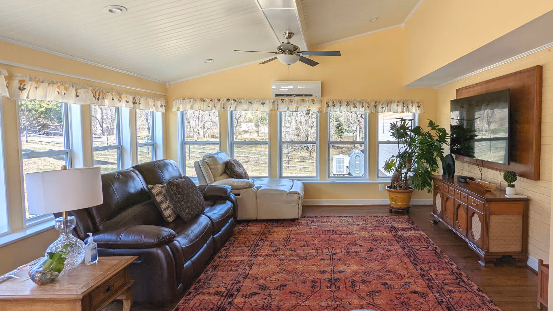 A bright living room with yellow walls and multiple windows letting in natural light. The room features a dark brown leather sofa with decorative pillows, a beige recliner chair, a large patterned area rug, a wooden side table with a lamp and hand sanitizer, a potted plant, and a wall-mounted flat-screen TV above a wooden cabinet.