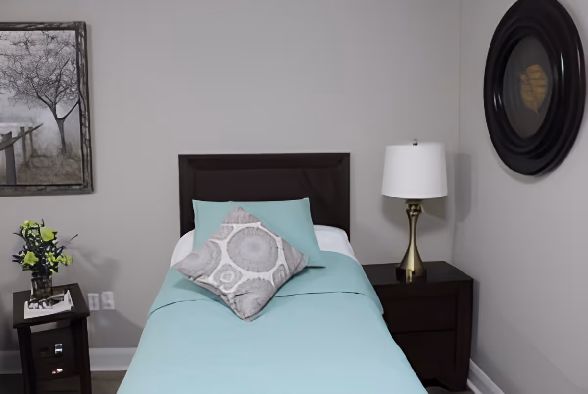 A small bedroom with a single bed covered in a light blue bedspread and a decorative pillow. There is a dark wooden headboard, a nightstand with a lamp on the right side, and a small table with a vase of flowers on the left side. The walls are light gray with a framed picture of a tree and a round black-framed mirror.