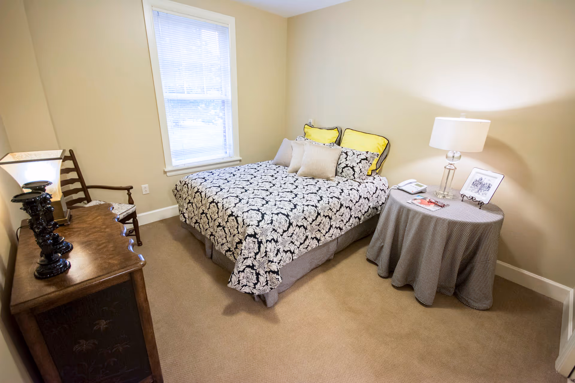 A small bedroom with a bed covered in a black and white patterned bedspread and yellow pillows. There is a round side table with a checkered tablecloth, a lamp, a phone, and a framed picture. A wooden dresser with decorative items and a chair are also visible. A window with blinds is on the wall behind the bed.