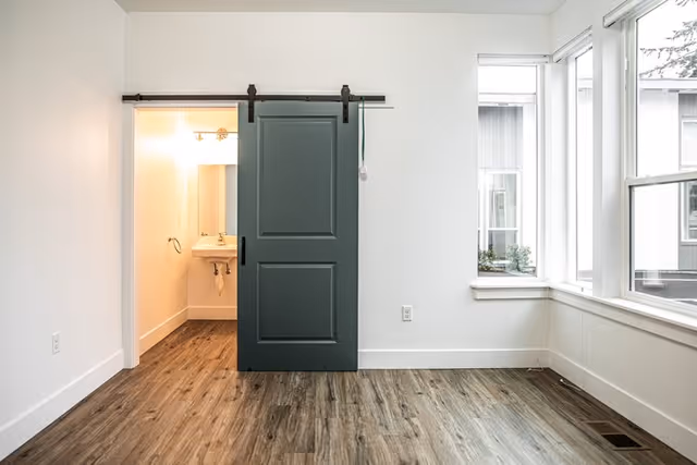 A bright room with wood flooring and white walls featuring a large window on the right side. A dark green sliding barn door is partially open, revealing a small bathroom with a white sink and mirror inside.
