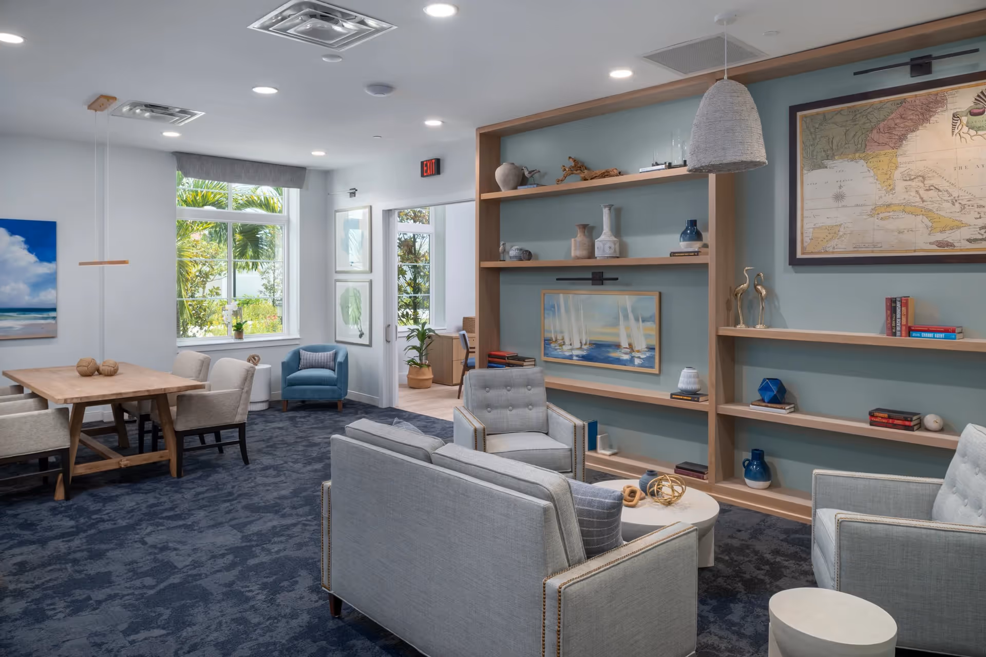 Bright community lounge with sofas and armchairs, built-in shelving, a dining table, and large windows.