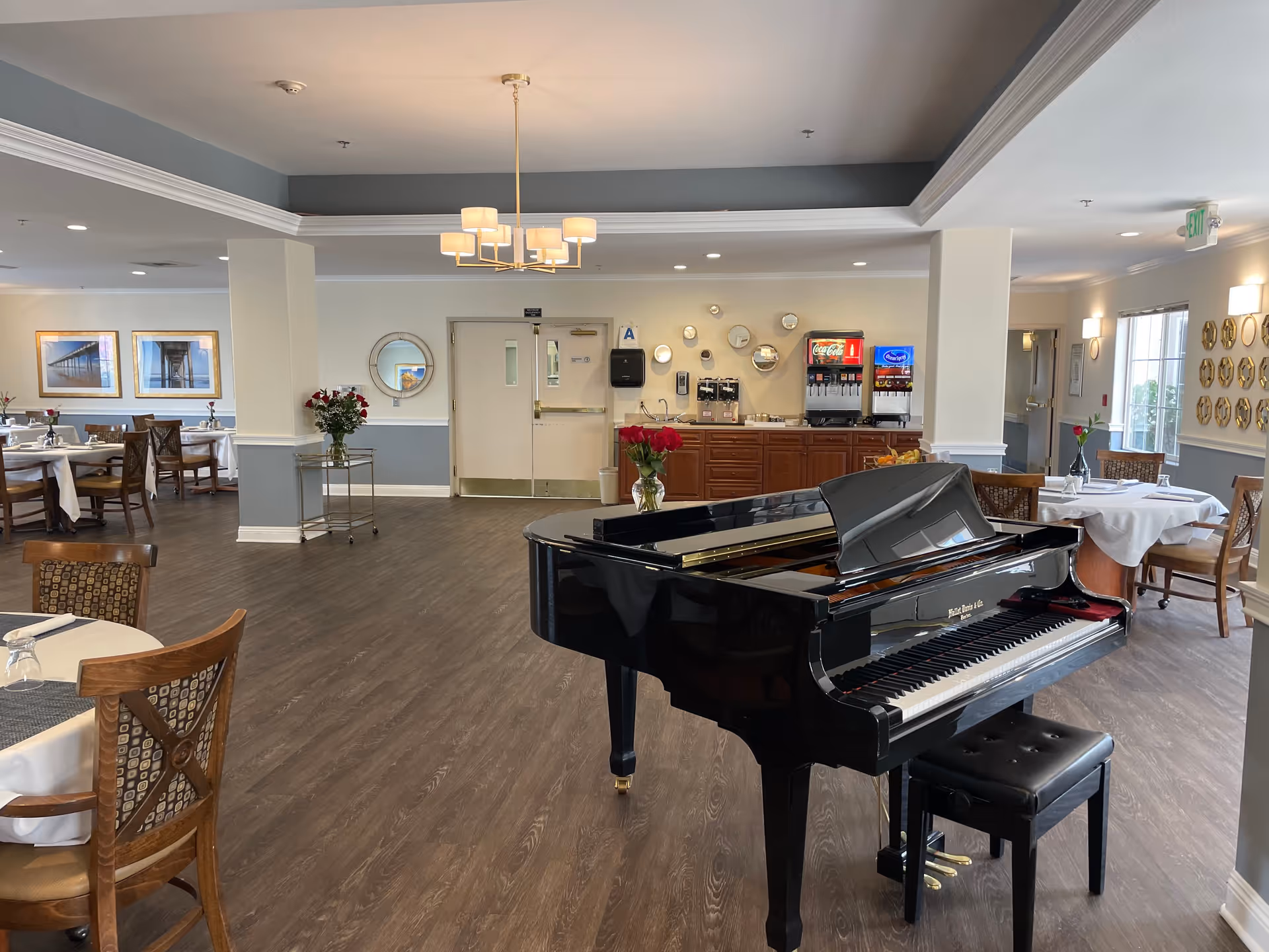 A spacious dining area with a black grand piano in the foreground. The room features wooden chairs and tables set with white tablecloths and glassware. On the back wall, there is a beverage station with soda dispensers and a coffee machine. The walls are decorated with framed pictures and round mirrors, and there are vases with red roses on some tables. The flooring is wood, and the ceiling has recessed lighting and a modern chandelier.