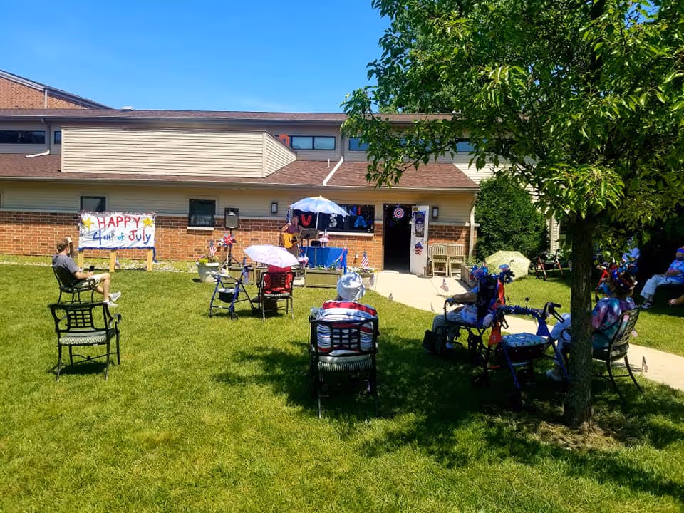 Outdoor gathering at The Pillars at Crystal Bay with several elderly people seated on chairs and walkers on a grassy lawn. A person is playing guitar under a white umbrella near the building. A colorful sign reads 'HAPPY 4th of July' attached to the building wall. The building is a single-story structure with brick and siding exterior. Trees provide some shade on the lawn.