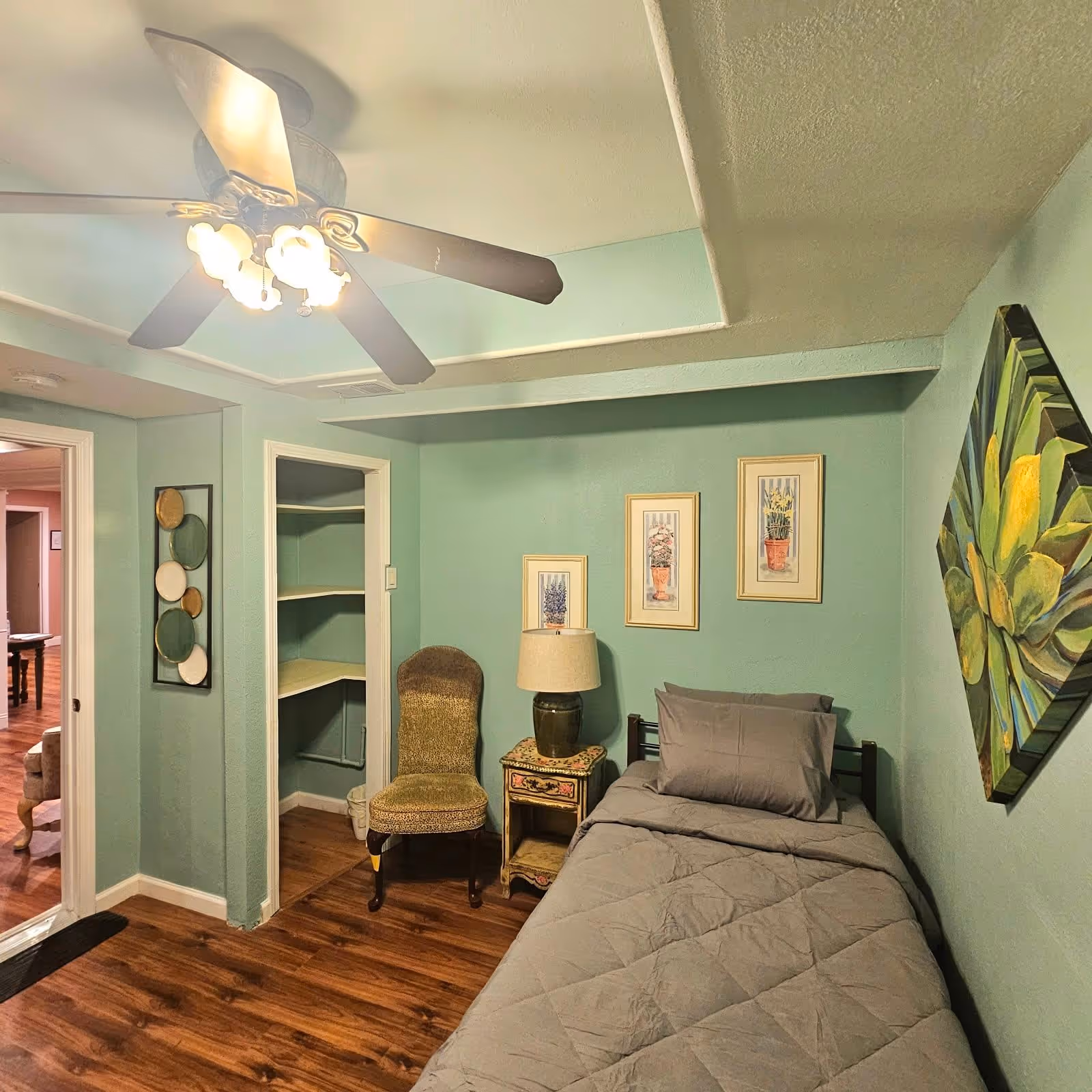 A small bedroom with light green walls and wood flooring. The room features a single bed with gray bedding, a vintage-style nightstand with a lamp, a green upholstered chair, and three framed floral prints on the wall above the nightstand. There is also a large painting of a succulent plant on the adjacent wall. A ceiling fan with lights is mounted on the ceiling, and an open doorway leads to another room with wood flooring.