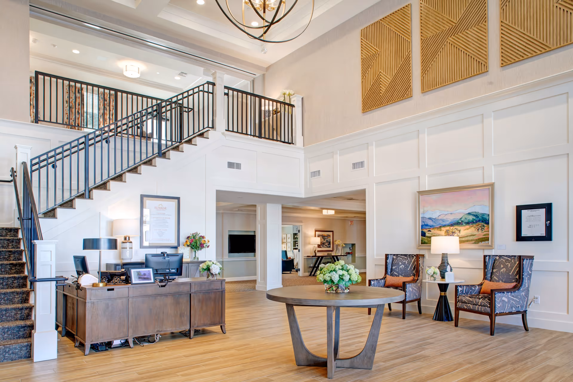 Bright and spacious senior living facility lobby with a wooden reception desk, two computer monitors, and a lamp. A staircase with black railings leads to an upper floor. A round wooden table with a flower arrangement is centered in the room. Two patterned armchairs with orange cushions flank a small side table with a lamp and decorative items. The walls are white with decorative paneling and artwork, including a landscape painting and framed documents.