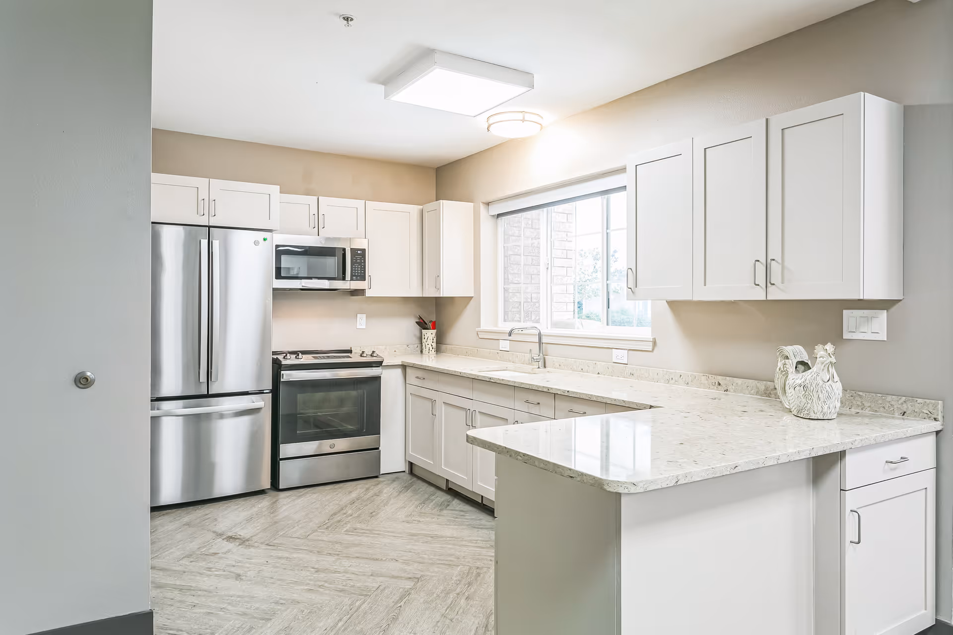 Bright modern kitchen with stainless steel refrigerator and stove, white cabinets, and a marble countertop peninsula beneath a window.