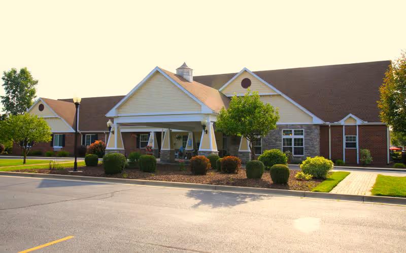 Exterior view of a single-story senior living facility building with a covered entrance, well-maintained landscaping including bushes and trees, and a paved parking area in front.
