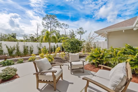 Outdoor patio area with cushioned wooden chairs arranged on a concrete surface, surrounded by landscaped garden beds with various green plants and small trees under a partly cloudy blue sky.