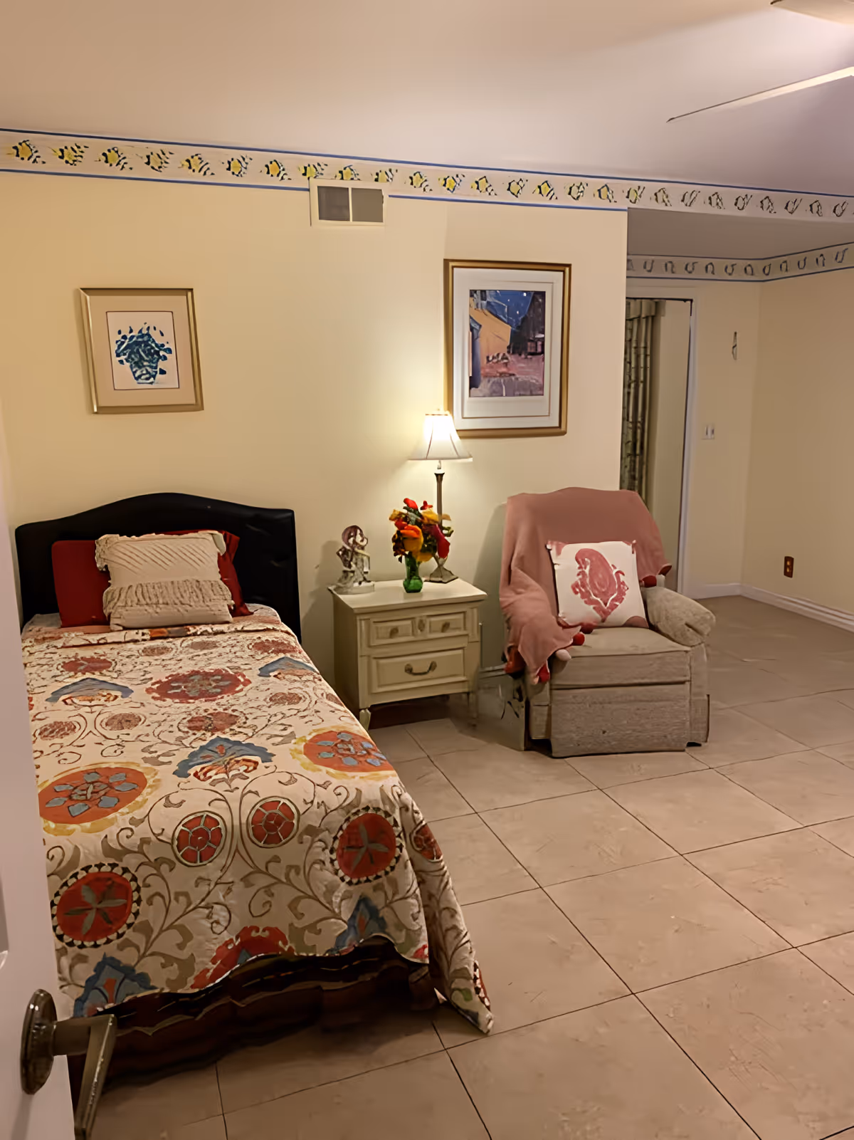 A cozy bedroom with a single bed covered in a colorful patterned quilt, a nightstand with a lamp and decorative items, and a comfortable armchair draped with a pink blanket and a decorative pillow. The walls are light yellow with a decorative border near the ceiling, and two framed pictures hang on the wall.