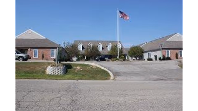 Exterior view of a single-story assisted living facility with a central building and two wings, an American flag on a flagpole in front, a driveway, and some trees and shrubs under a clear blue sky.