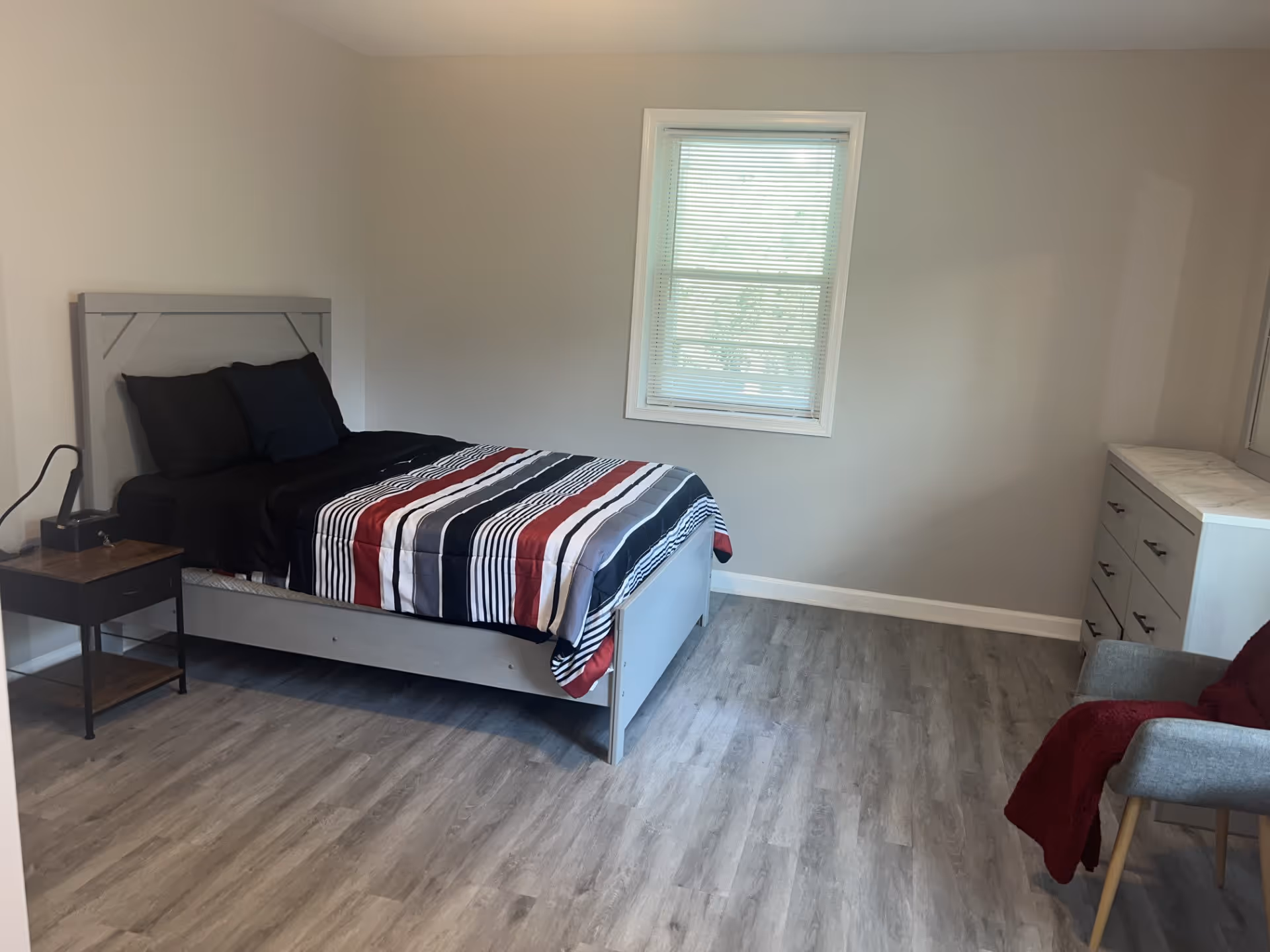 A simple bedroom with a single bed featuring a striped comforter in black, white, and red. There is a small wooden nightstand with a lamp on the left side of the bed. A window with blinds is centered on the wall behind the bed. On the right side of the room, there is a white dresser with a marble-like top and a gray chair draped with a red blanket. The floor is covered with light wood laminate.