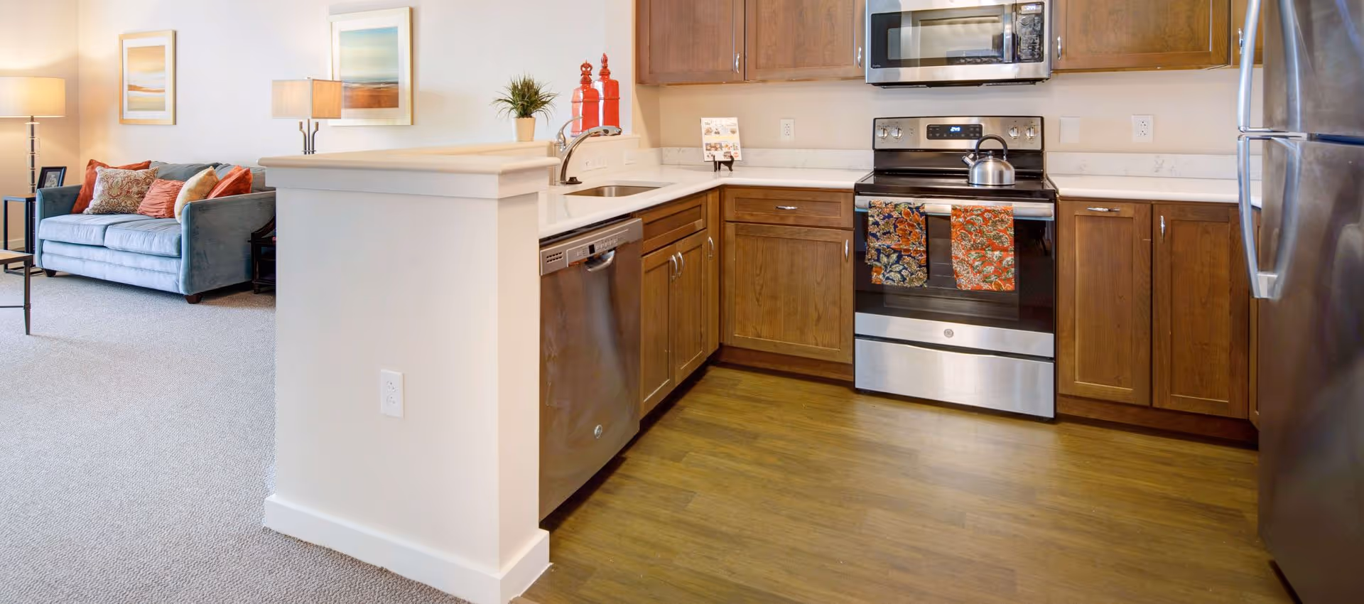 Open kitchen with stainless steel refrigerator, stove and dishwasher, wooden cabinets, and a nearby living area with a couch.