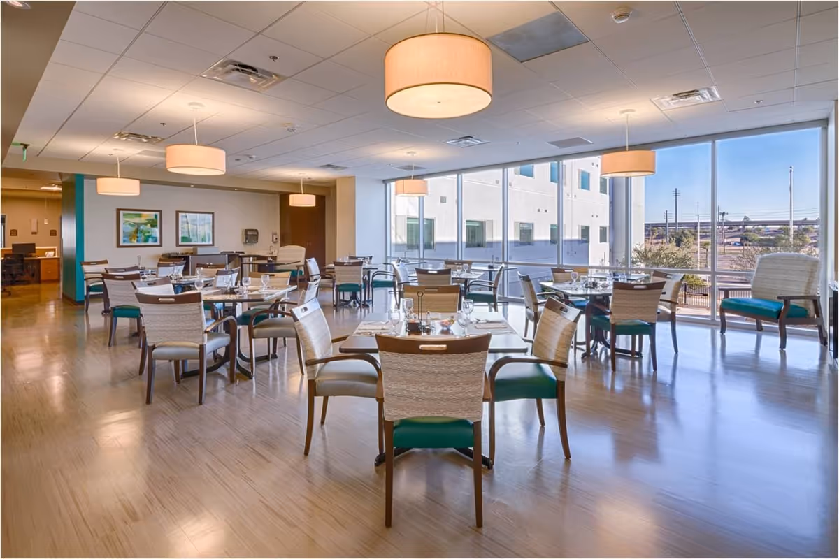 A spacious dining room with multiple round tables set with glasses and napkins. The room has large floor-to-ceiling windows allowing natural light to fill the space, and modern hanging light fixtures. Chairs with wooden frames and green cushions surround the tables. Artwork is displayed on the far wall, and the floor is a light wood finish.