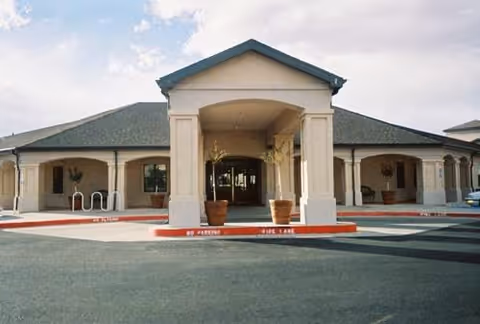 Front entrance of a single-story senior living facility with a covered porte-cochere, columns, and potted plants.