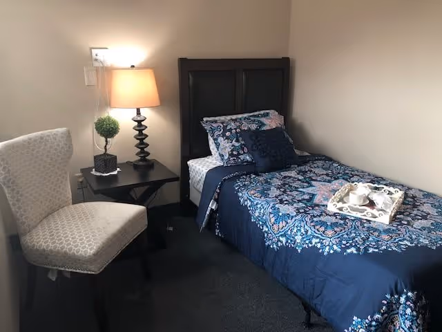 A small bedroom with a single bed covered in a blue patterned bedspread and matching pillows. Next to the bed is a black nightstand with a lamp and a small decorative plant. A light-colored upholstered chair is positioned near the nightstand. The walls are plain and light-colored.