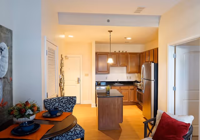 View of a cozy kitchen and dining area in a senior living facility. The kitchen features wooden cabinets, a stainless steel refrigerator, a small island with a plant on top, and a hanging light fixture. Adjacent to the kitchen is a round dining table set with two blue patterned chairs, orange placemats, and blue dishes. A white door is visible in the background, and a comfortable chair with a red pillow is partially visible on the right side.
