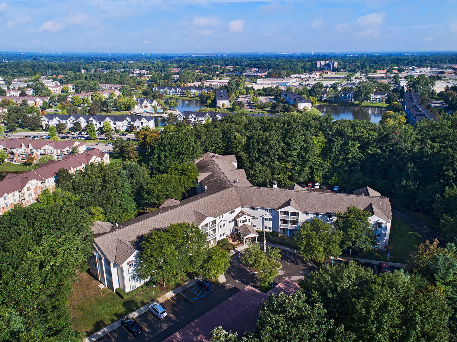 Aerial view of a curved senior living building surrounded by trees, parking areas and nearby ponds and houses.