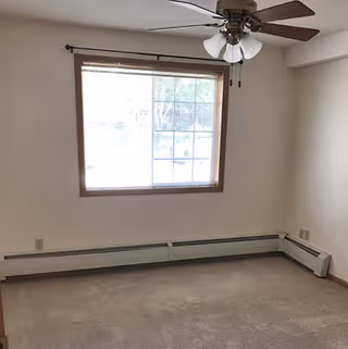 Empty room with beige carpet, white walls, a large window with blinds, a ceiling fan with lights, and baseboard heating along two walls.