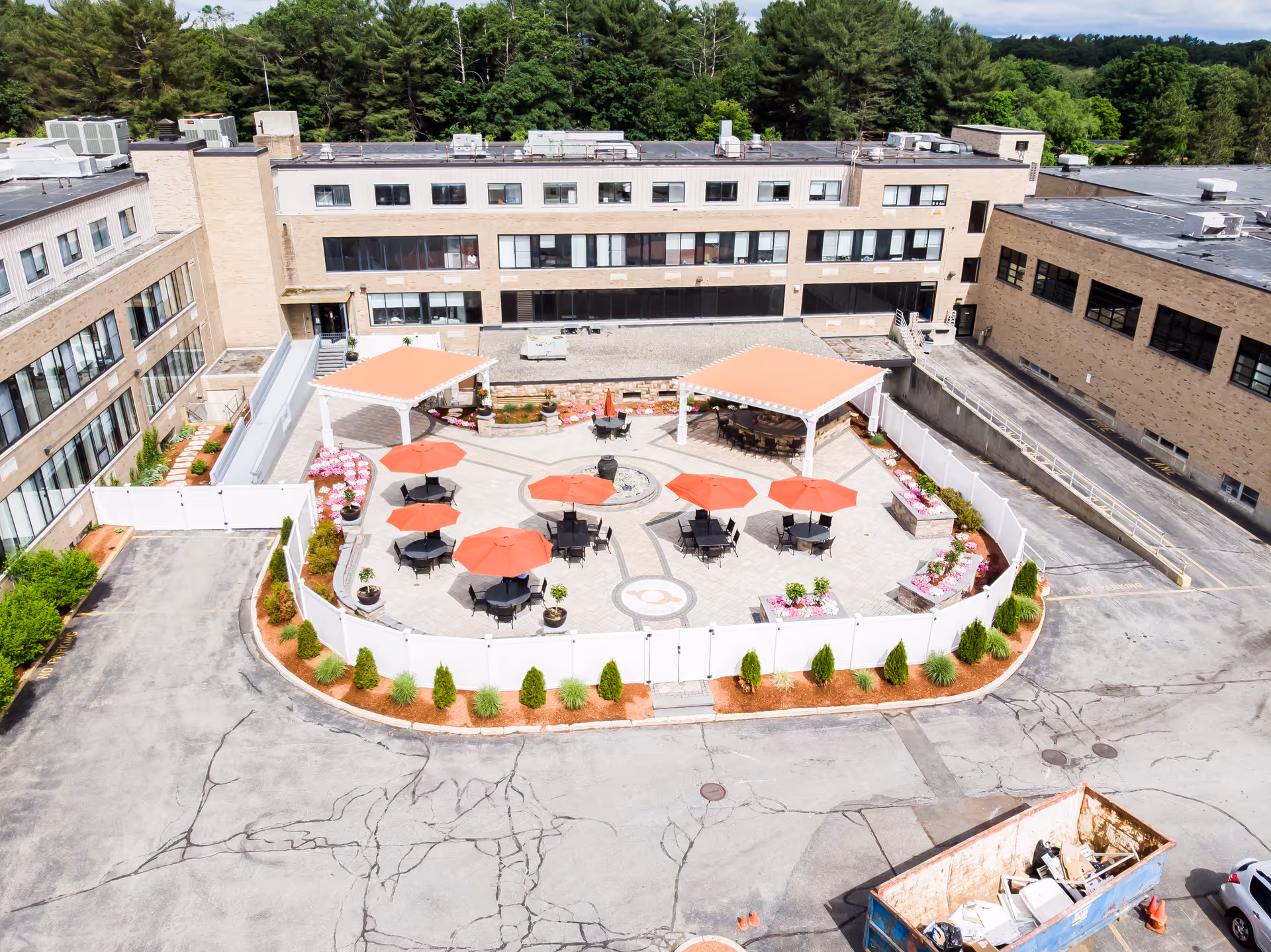 Aerial view of an outdoor patio area at CareOne at Concord, featuring multiple black tables with red umbrellas, two covered pergolas, surrounding flower beds, and a white fence enclosing the space. The patio is situated within a U-shaped building with beige brick walls and multiple windows, surrounded by a paved parking area and some greenery in the background.