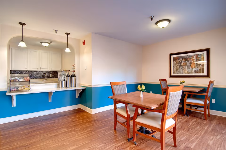 A cozy dining area in a senior living facility with two wooden tables, each surrounded by four cushioned chairs. The walls are painted beige with a teal wainscoting. A framed artwork hangs on the wall. To the left, there is a counter with a pass-through window to a kitchen area, featuring white cabinets, a mosaic tile backsplash, and coffee dispensers. The floor is wood laminate, and the room is softly lit by ceiling lights and pendant lamps.