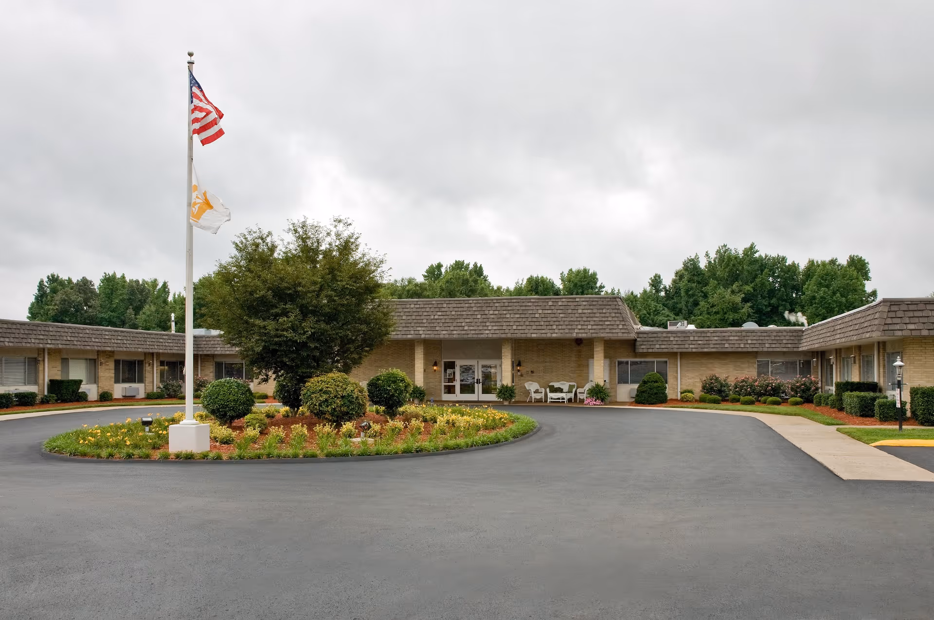 Single-story care facility entrance with a circular driveway, flagpole, landscaped island, and seating on the front porch.