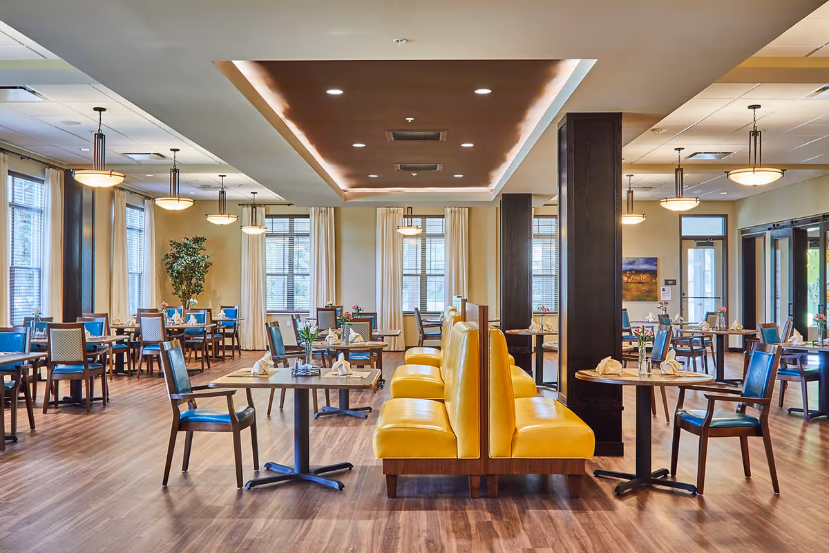 A spacious dining room with wooden floors, yellow cushioned booth seating in the center, and multiple tables with blue chairs arranged around the room. The room is well-lit with large windows covered by light curtains and ceiling lights hanging from the ceiling. The tables are set with napkins and small flower vases.