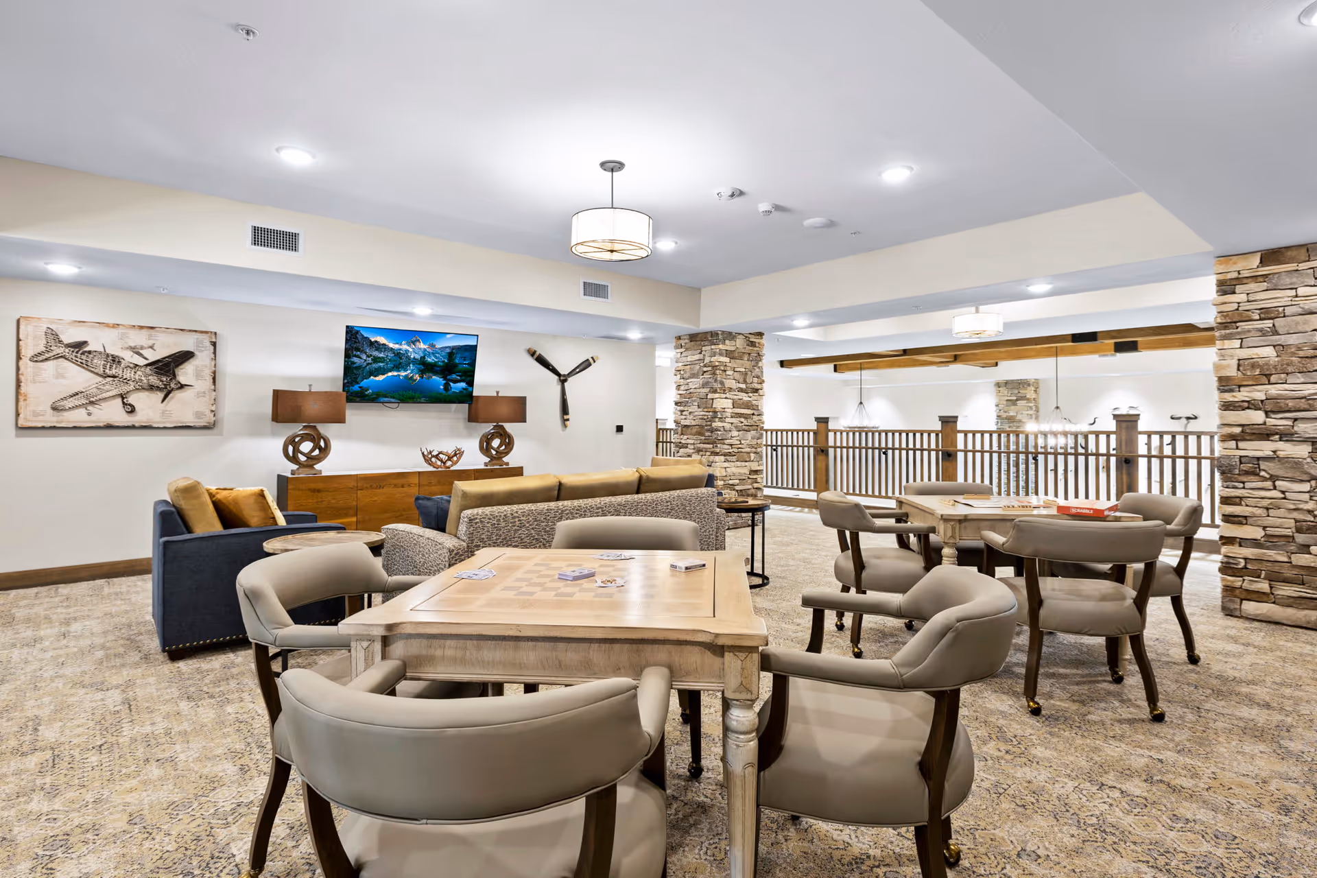 Spacious senior living common area with game tables, lounge seating, stone pillars and a wall-mounted TV.