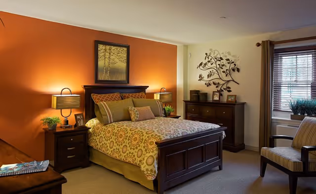 Cozy furnished bedroom with a wooden bed, patterned bedding, nightstands with lamps, dresser, wall art, and a window with blinds.