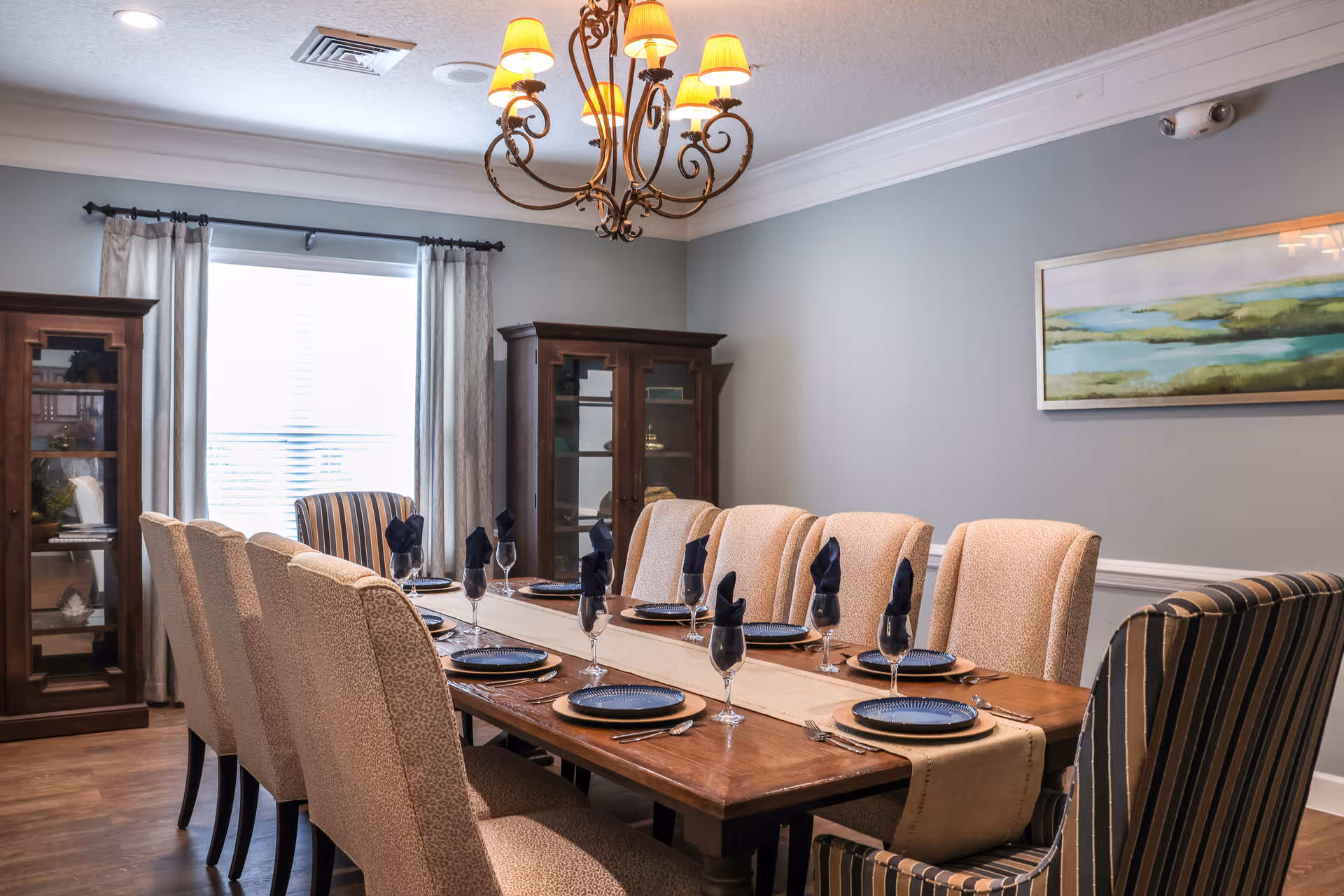 A formal dining room with a long wooden table set for eight people. The table has a beige runner, blue plates, wine glasses with black napkins, and silverware. The room features beige upholstered chairs and two striped armchairs at the ends of the table. There are two wooden display cabinets against the wall, a window with light curtains, a chandelier with yellow lampshades hanging from the ceiling, and a landscape painting on the wall.