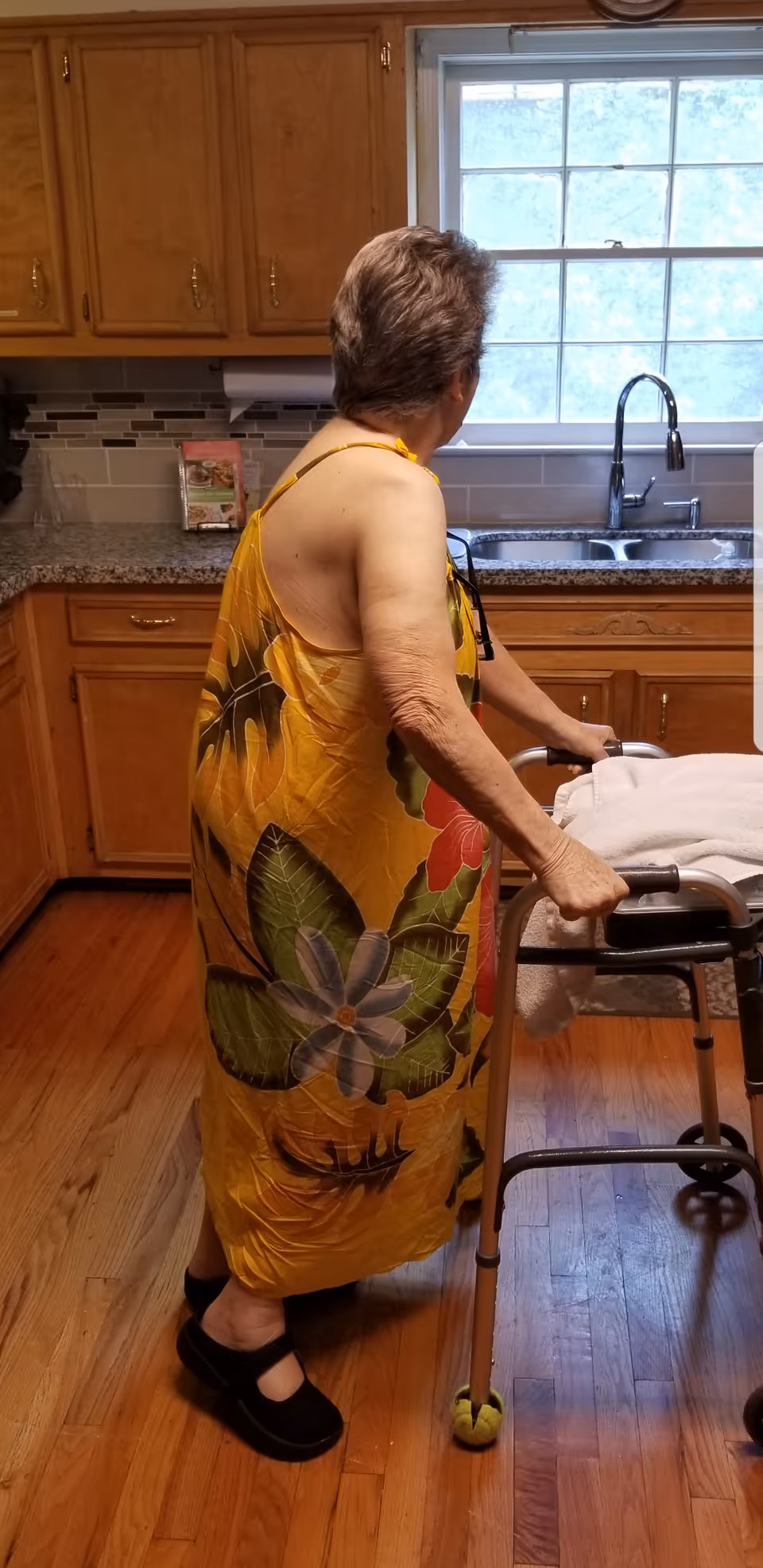 A person using a walker stands in a kitchen near a sink and wooden cabinets.
