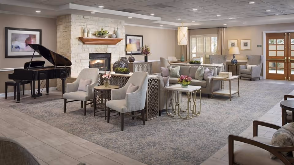 A spacious and elegantly furnished senior living common area featuring a grand piano, a stone fireplace with a wooden mantle, multiple armchairs and sofas with cushions, side tables with flower arrangements, framed artwork on the walls, large windows with curtains, and a wooden door with glass panels.