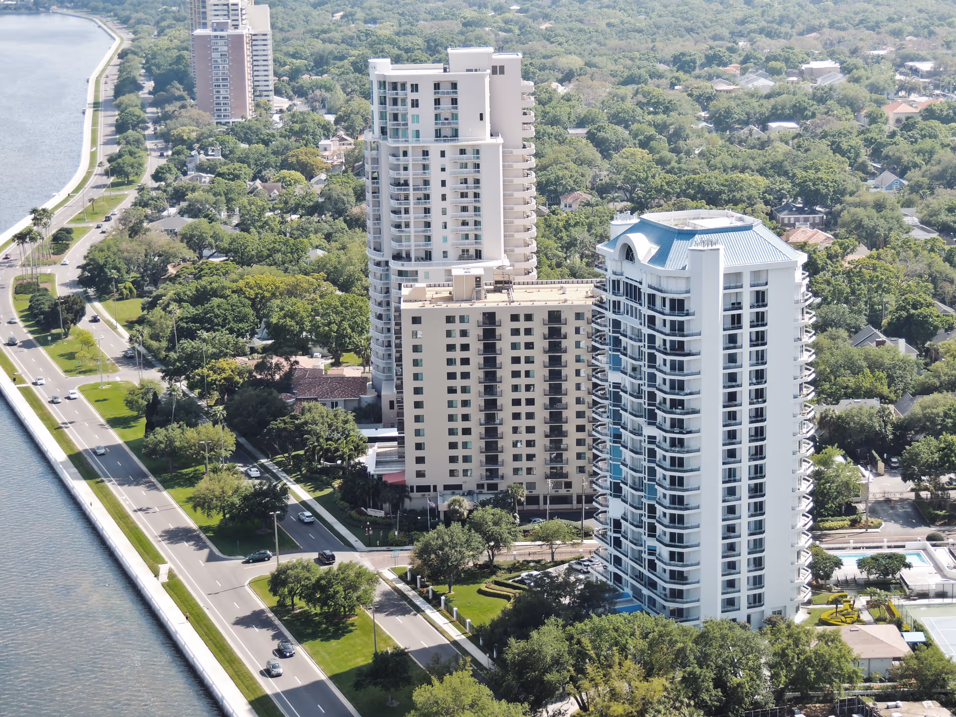 Aerial view of several tall residential buildings surrounded by trees and greenery, with a road and waterway running parallel on the left side. The buildings are part of an urban neighborhood with a mix of high-rise and low-rise structures.