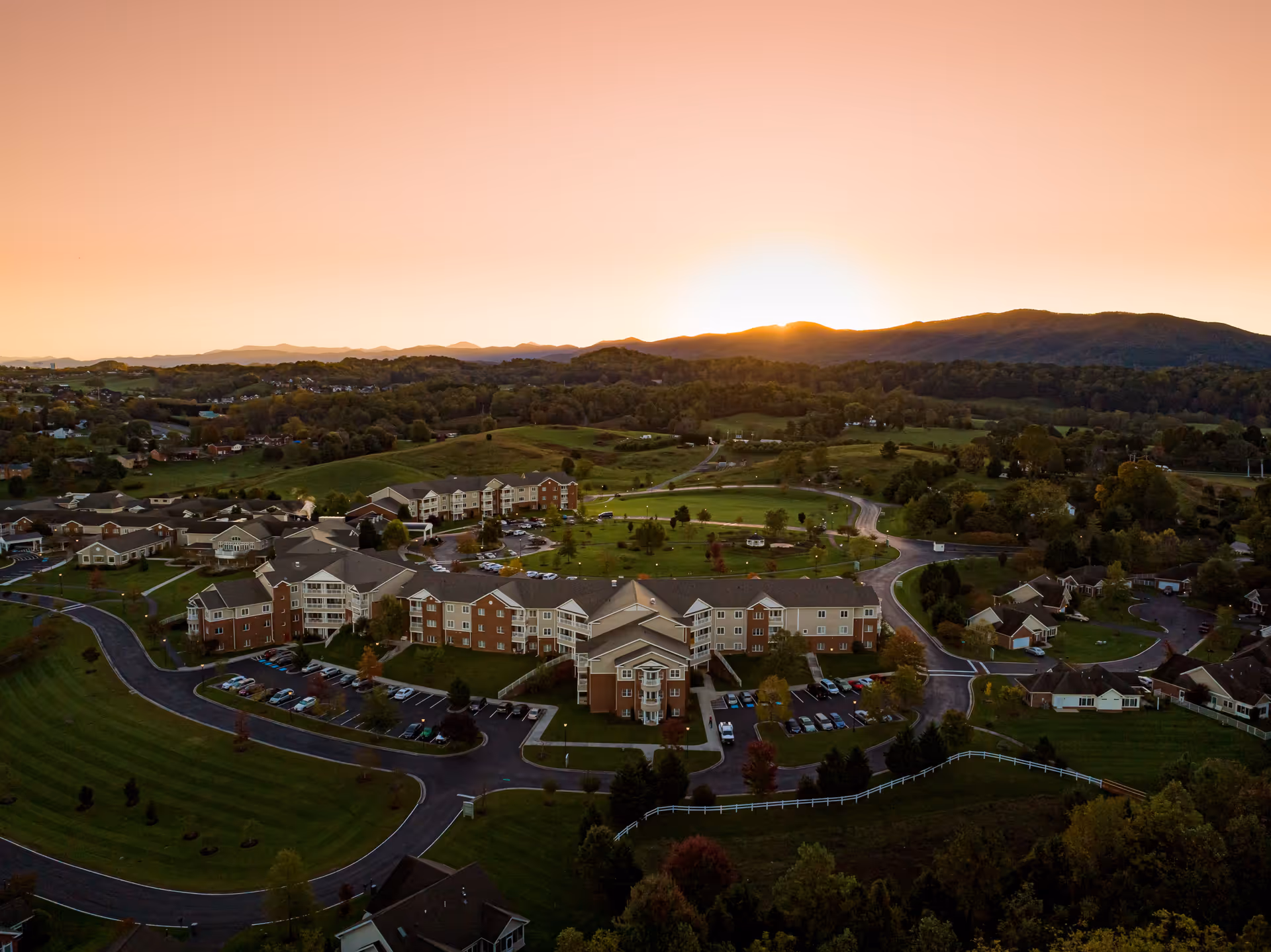 Aerial view of The Glebe Retirement Community campus with multiple brick residential buildings, parking lots, winding roads and surrounding green hills at sunset.