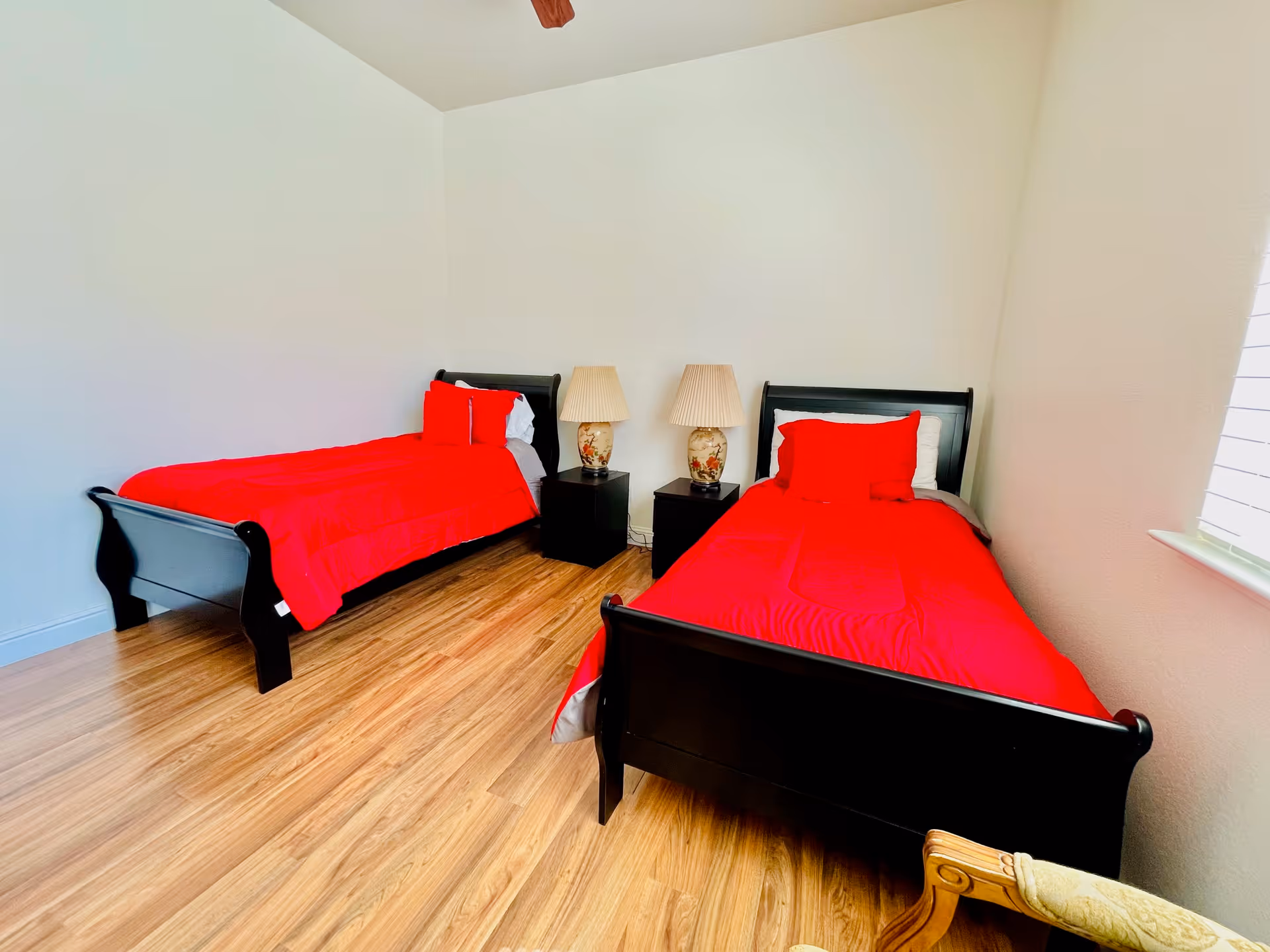 A bedroom with two single black wooden beds, each made with bright red bedding and pillows. Between the beds are two dark nightstands, each with a decorative lamp. The room has light-colored walls, a wooden floor, a ceiling fan, and a window with blinds on the right side.