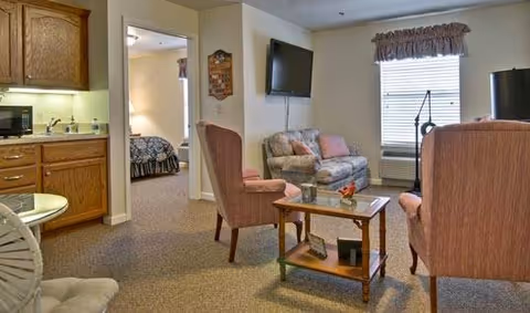 A cozy senior living facility interior featuring a small living room area with two upholstered chairs, a loveseat, a glass coffee table, and a wall-mounted TV. To the left, there is a kitchenette with wooden cabinets, a microwave, and a sink. A doorway leads to a bedroom with a bed visible in the background. The room is carpeted and has a window with a valance, an air conditioning unit, and a floor lamp.