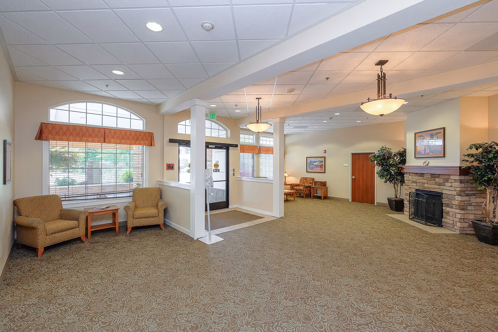 A spacious and well-lit common area with beige walls and patterned carpet. The room features two armchairs with a small wooden table between them near large windows with orange valances. There is a stone fireplace with a mantel on the right side, flanked by potted plants. The ceiling has recessed lighting and two hanging light fixtures. A door and framed artwork are visible on the far wall.