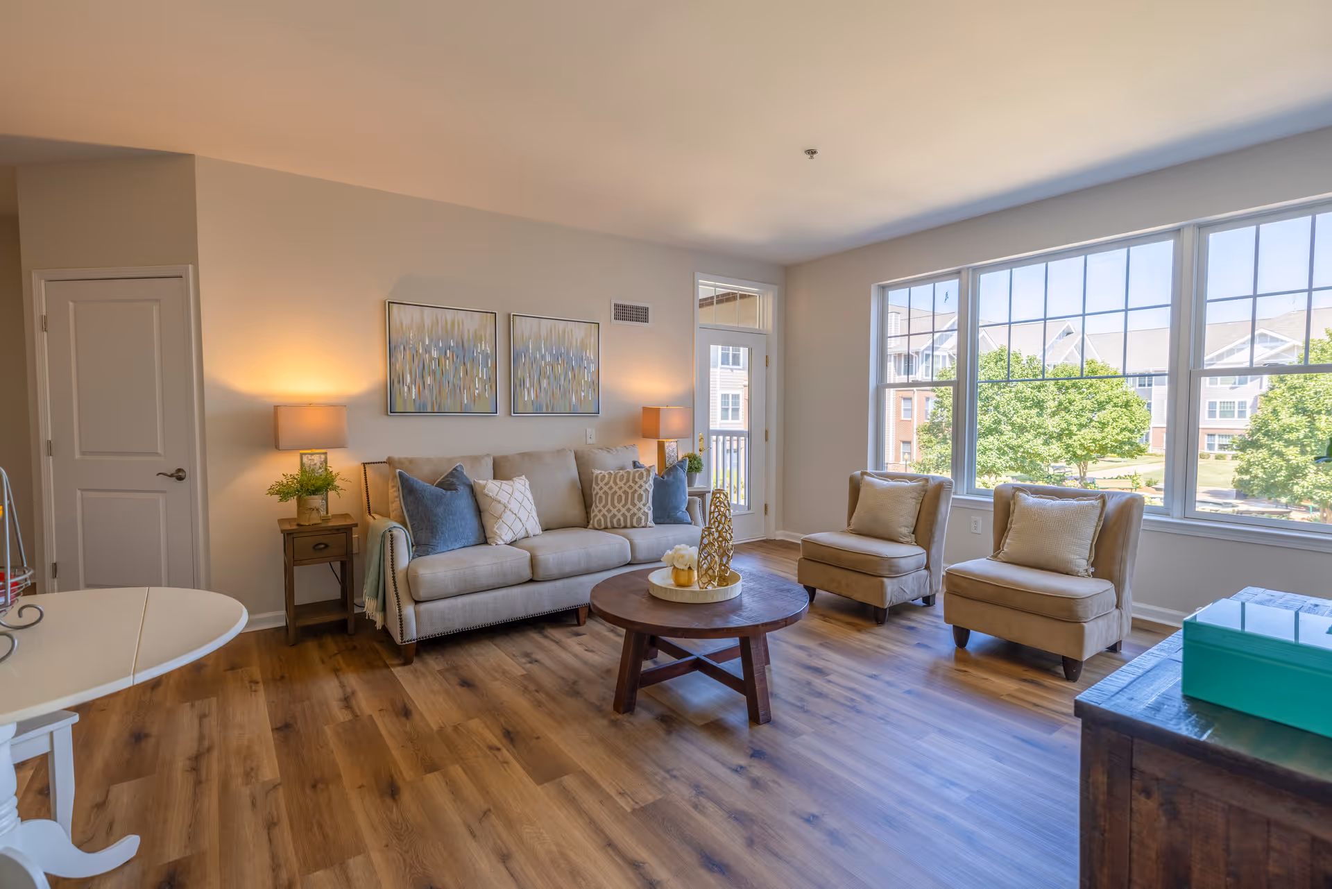 Bright furnished living room with a sofa, two armchairs, a round coffee table, and large windows overlooking greenery.