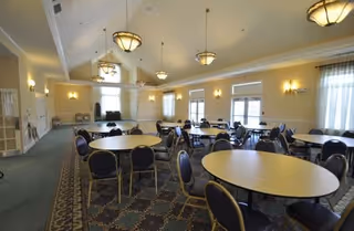 Large dining/event room with multiple round tables and chairs, chandeliers, and windows along the walls.
