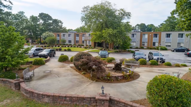 Outdoor view of Millennium Post Acute Rehabilitation facility showing a parking lot with several cars, landscaped garden areas with bushes and trees, benches, and a two-story building in the background.