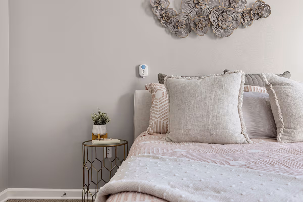 A neatly made bed with multiple beige and patterned pillows against a light gray wall. A small round side table with a glass top holds a small potted plant and a book. Above the bed is a decorative wall art piece featuring metallic flowers.