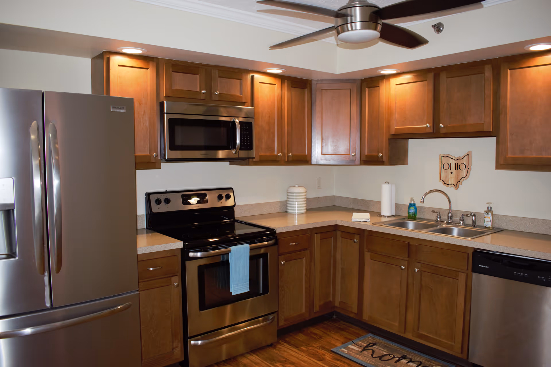 A modern kitchen with wooden cabinets, stainless steel refrigerator, oven, microwave, and dishwasher. The kitchen features a double sink with a faucet, a paper towel holder, and a decorative Ohio state wall hanging. The floor is wooden, and there is a ceiling fan with lights above.