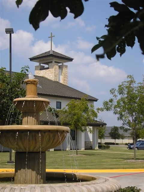 Outdoor view of a fountain in front of a chapel-like building with a cross-topped tower and landscaped grounds.