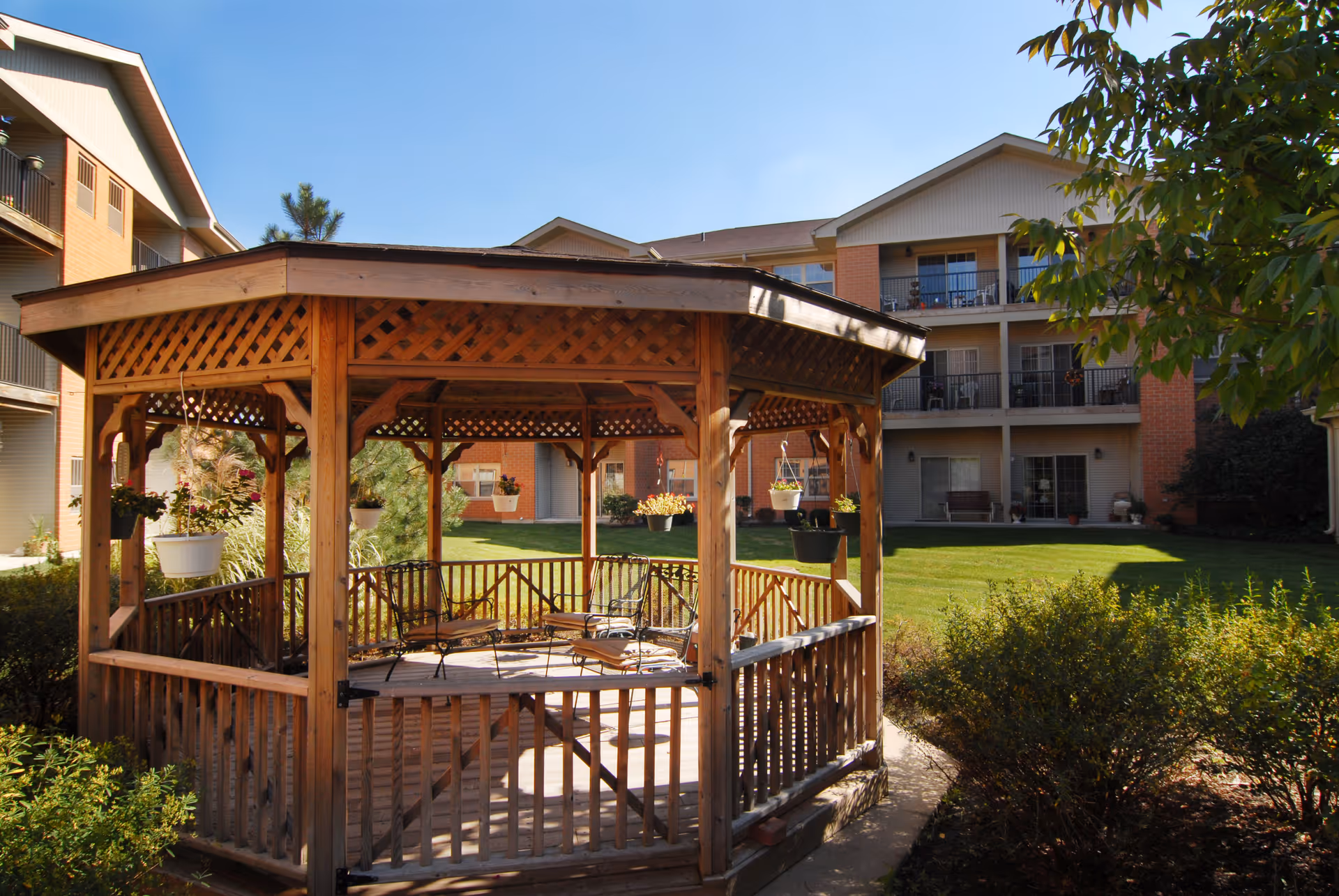 Wooden gazebo with chairs in a grassy courtyard in front of a multi-story senior living building.