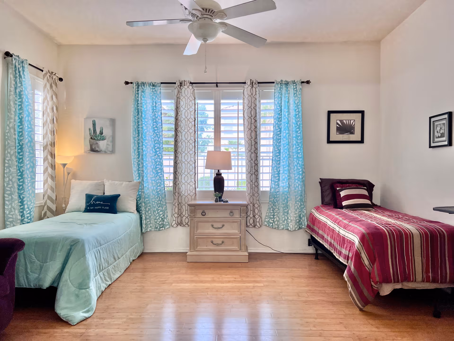 A bright bedroom with two single beds on either side of a window with blue and beige patterned curtains. The left bed has a light blue comforter and a decorative pillow that says 'home is my happy place.' The right bed has a red and beige striped comforter with matching pillows. A wooden nightstand with a lamp is positioned between the beds. The room has light-colored walls, wooden flooring, a ceiling fan, and framed artwork on the walls.