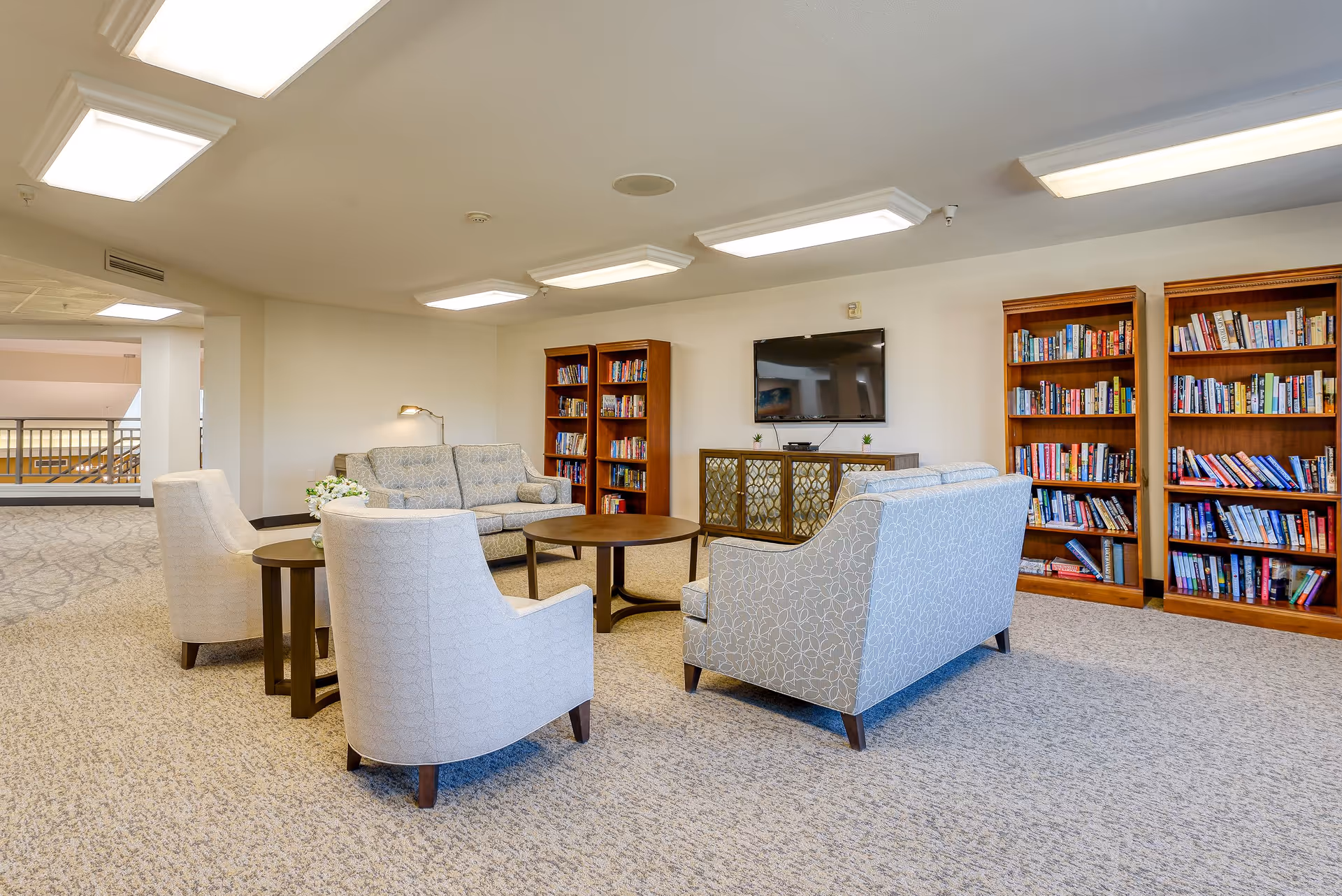 A cozy senior living common area with two armchairs and two sofas arranged around a round wooden coffee table. Behind the seating area are three wooden bookshelves filled with books and a flat-screen TV mounted on the wall above a decorative cabinet. The room has beige carpet and bright ceiling lights.