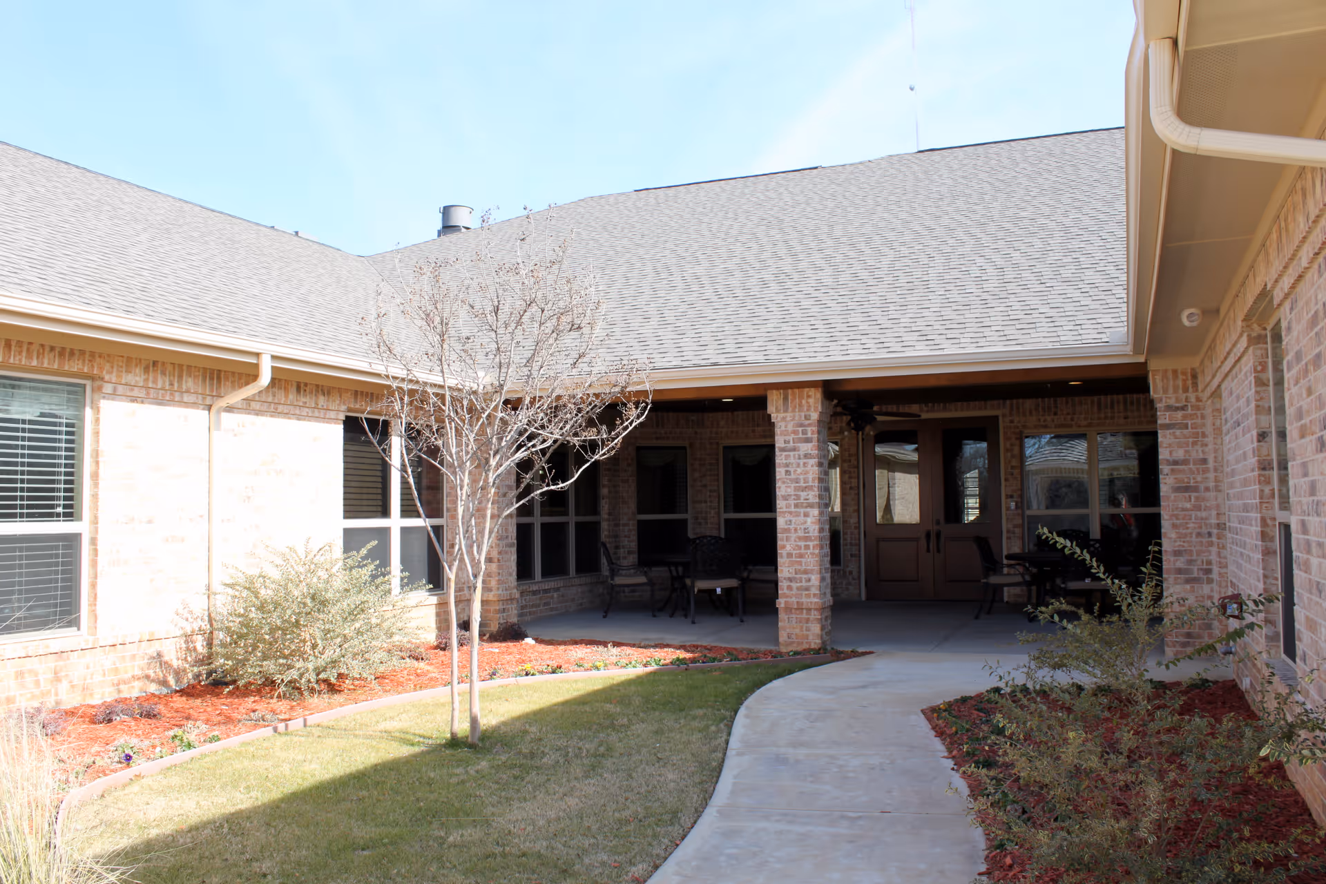 Outdoor courtyard area of a brick building with a covered patio featuring chairs and tables. There is a small tree and landscaped garden beds with mulch along the walkway leading to the patio entrance.