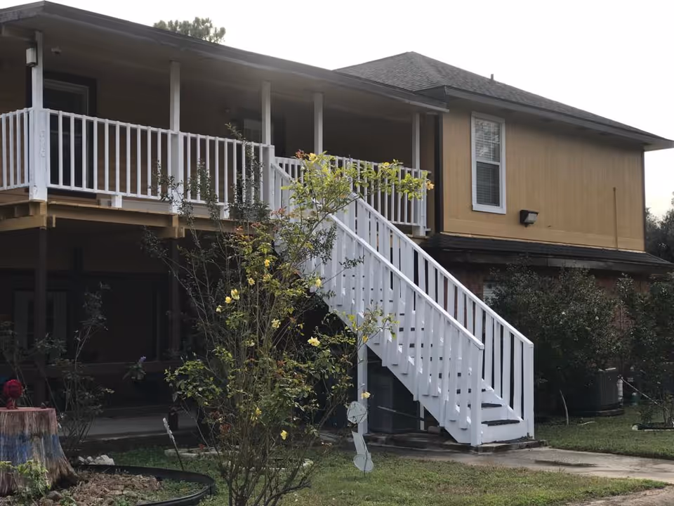 Exterior view of a two-story building with a white wooden staircase leading to a second-floor porch with white railings. There are bushes and a tree with yellow flowers in the foreground, and the building has a beige upper exterior with a dark roof.