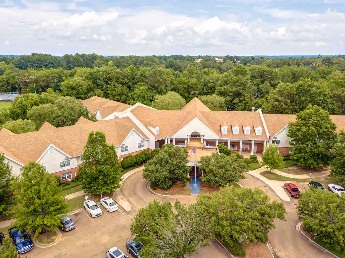 Aerial view of Ridgeland Place senior living building showing the front entrance, circular driveway, parked cars, and surrounding trees.
