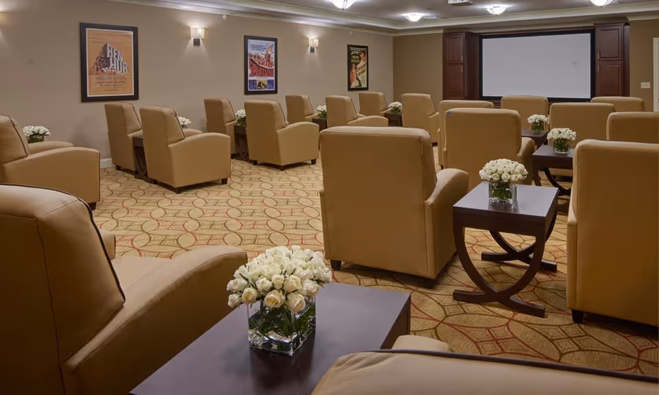 A cozy movie or presentation room with multiple tan leather armchairs arranged in rows facing a large screen. Each armchair is paired with a small dark wooden side table holding a vase of white roses. The walls are decorated with framed vintage movie posters and soft wall-mounted lights provide warm illumination.